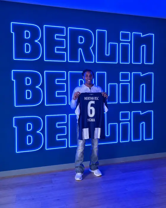 Jermaine Ngwa hält sein Trikot von Hertha BSC vor einer Leuchtschrift mit dem Schriftzug Berlin